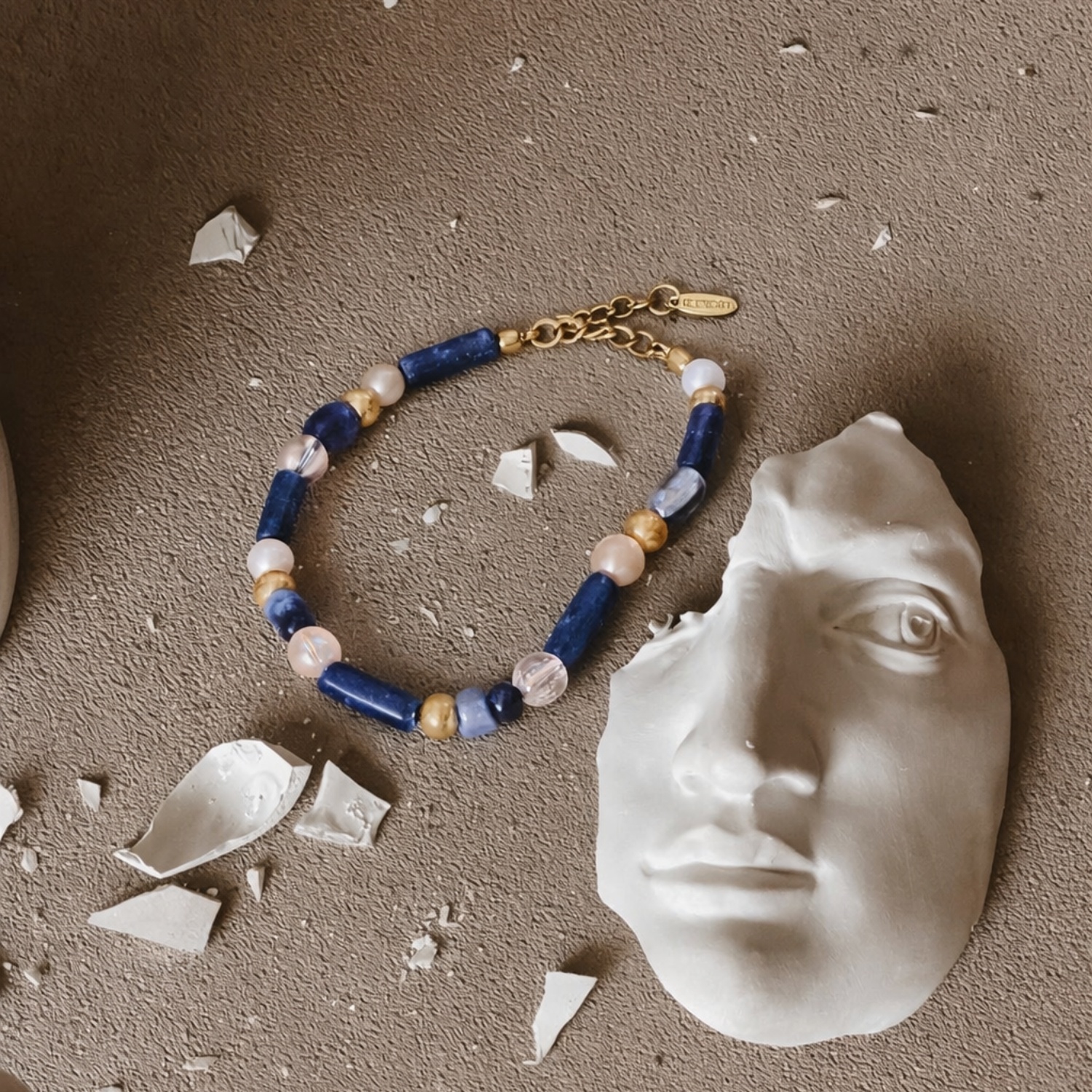 Athens Bracelet en composició bodegó amb una màscara clàssica blanca i fragments de ceràmica, polsera de gemstones blaus, blancs i accents daurats.
