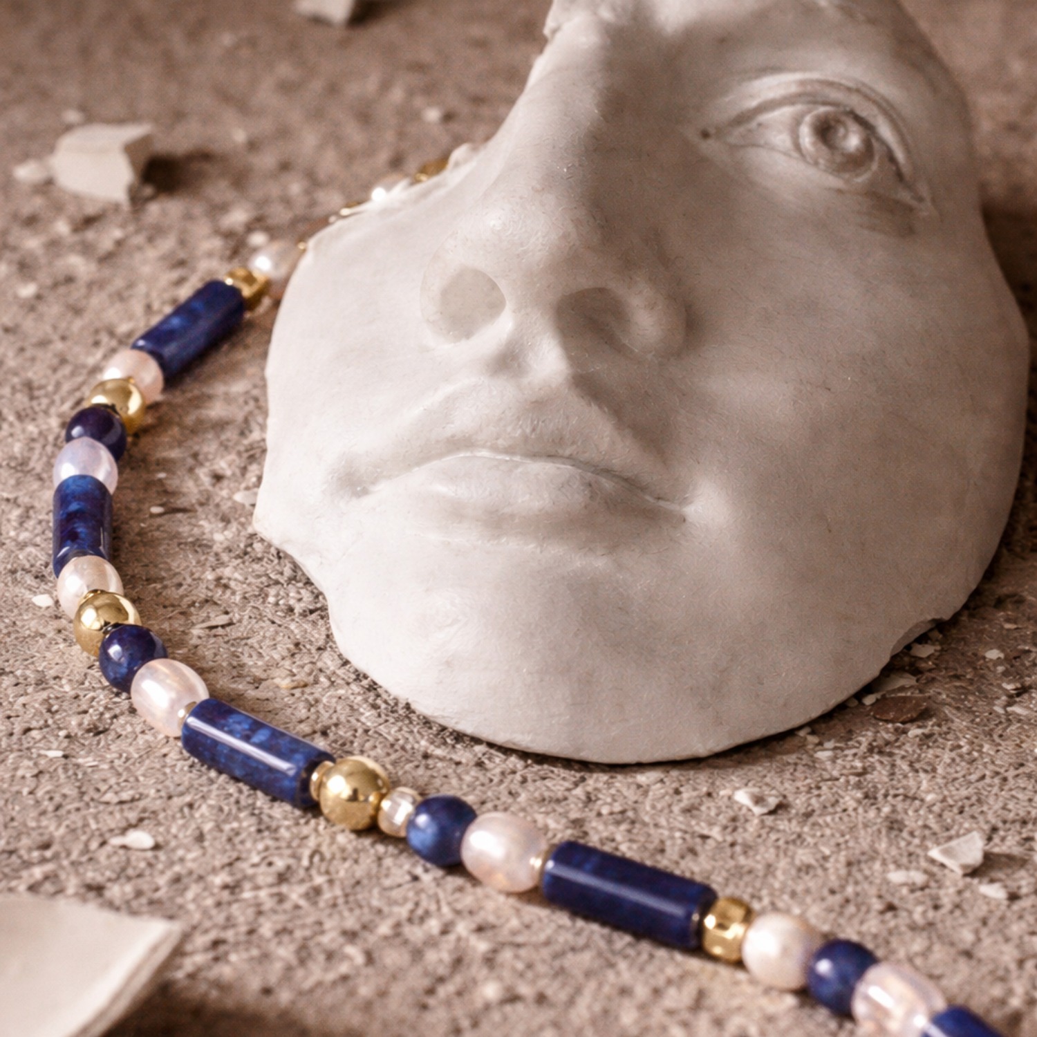 Athens Necklace in a still life composition with blue gemstone beads, white details, and gold accents beside a sculptural face.